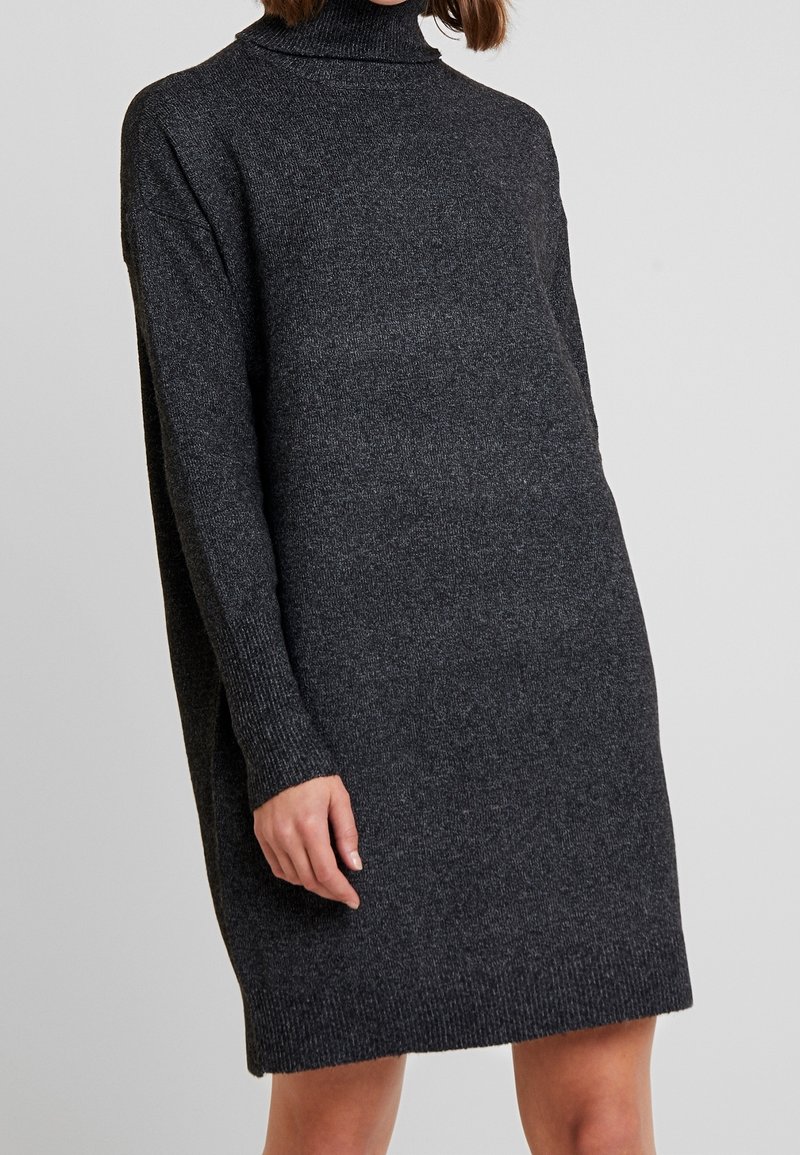 Robe-pull en maille gris foncé à manches longues avec col roulé portée par une personne devant un fond uni gris clair.