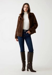Giacca in pelliccia sintetica marrone, dolcevita bianco, jeans scuri in denim e stivali marroni in pelle al ginocchio con dettaglio di cerniera dorata.