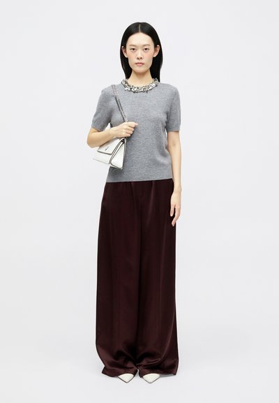 Szary sweter z krótkim rękawem, zdobiony na dekolcie, zestawiony z szerokimi burgundowymi spodniami. Modelka trzyma małą białą torebkę z srebrnymi elementami.
