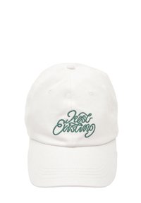 Casquette en coton blanc avec un texte brodé vert disant "Just Existing", dotée d'une visière incurvée et d'un design à six panneaux avec des trous de ventilation.