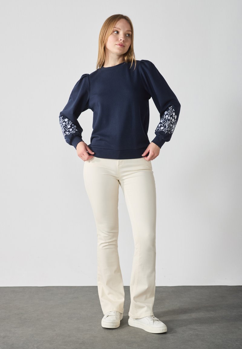 Donkerblauwe sweatshirt met poefmouwen, versierd met witte bloemenborduursels op de manchetten. Gecombineerd met crèmekleurige broek en witte sneakers.
