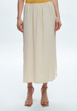 Frau trägt einen gelb-weiß vertikal gestreiften Midirock, kombiniert mit beigen Sandaletten mit hohen Absätzen, steht vor weißem Hintergrund.