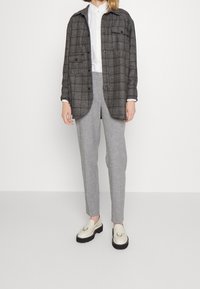 Grijs geruit overhemd jasje met twee voorzakken, gedragen over een wit overhemd met kraag, gecombineerd met lichtgrijze broek en crèmekleurige loafers met zwarte zolen.