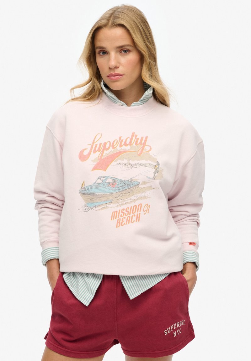Lichtroze sweatshirt met grafische print van een boot en de tekst "Superdry Mission CA Beach". Gepaard met bordeauxrode shorts en een gestreepte kraagshirt.