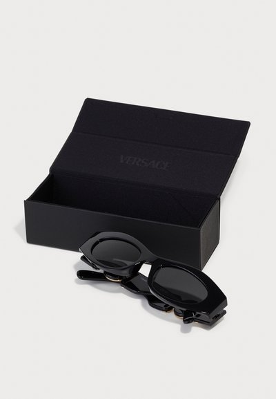 Des lunettes de soleil noires au design angulaire unique reposant sur une boîte Versace noire. Elles possèdent des verres sombres et des montures brillantes avec des accents dorés.