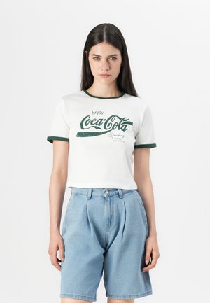 Balta trumpa marškinėliai su žaliais akcentais ir Coca-Cola logotipu. Pora su aukštu juosmeniu šviesiai mėlynomis džinsinėmis šortais. Lygi audinio tekstūra.