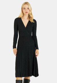 Schwarzes, gefaltetes Kleid mit langen Ärmeln, tiefem V-Ausschnitt, strukturiertem Material und subtiler Schimmer. Knielang, tailliert und weit auslaufend.