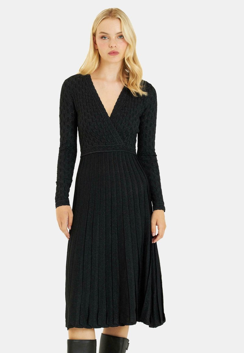 Schwarzes, gefaltetes Kleid mit langen Ärmeln, tiefem V-Ausschnitt, strukturiertem Material und subtiler Schimmer. Knielang, tailliert und weit auslaufend.