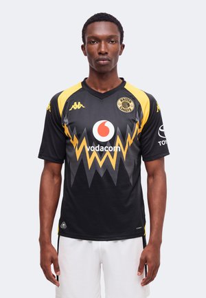 Homme debout portant un maillot de football Kaizer Chiefs noir et jaune avec un short blanc sur fond blanc uni.