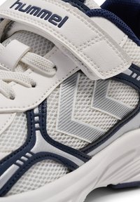Witte sportschoen met marineblauwe accenten, mesh materiaal, gestikte details, klittenbandsluiting en zilveren geometrische patronen aan de zijkanten.