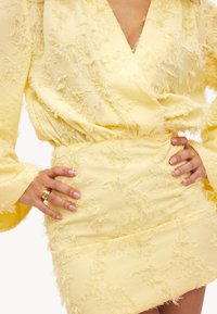 Robe jaune en tissu texturé et effiloché, avec un décolleté en V profond et des manches longues. La jupe est ajustée avec une taille lisse.