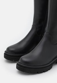 Black rubber knee-high boots with a smooth finish, featuring a side zipper, rounded toe, and a textured, chunky sole for grip.