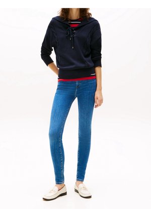 Femme portant un pull bleu marine avec un col lacé, une chemise rayée en dessous, un jean skinny bleu et des mocassins blancs, debout avec une main dans le dos.