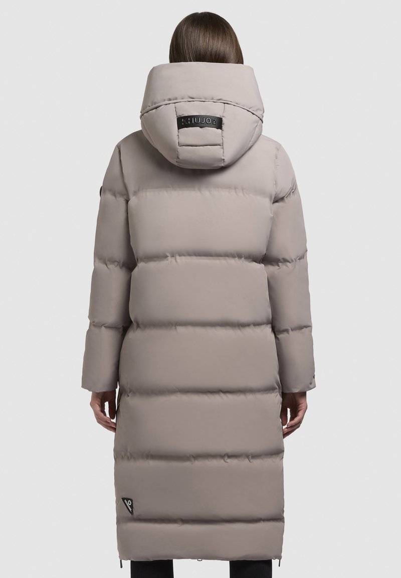 Manteau rembourré long de couleur gris clair, avec une grande capuche, des coussins horizontaux distincts et un patch logo au dos. Ouvertures zippées sur les côtés à l'ourlet.