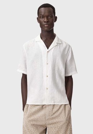 Ung mand iført en hvid kortærmet skjorte med knapper og beige bukser med mønster, stående med hænderne i lommerne mod en ensfarvet baggrund.