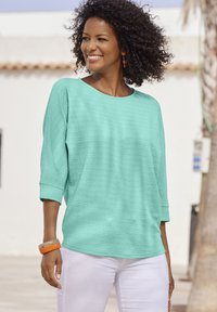Mintgrünes, strukturiertes Oberteil mit Rundhalsausschnitt und Dreiviertelärmeln, kombiniert mit weißen Hosen und einem orangen Accessoire.