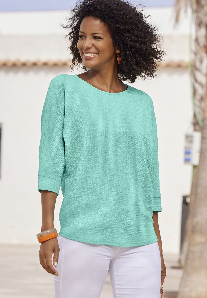 Mintgrünes, strukturiertes Oberteil mit Rundhalsausschnitt und Dreiviertelärmeln, kombiniert mit weißen Hosen und einem orangen Accessoire.