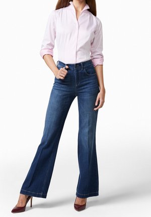 Kvinde iført en lyserød skjorte med knapper og op-rullede ærmer, høj talje med vidde blå jeans og vinrøde højhælede sko.