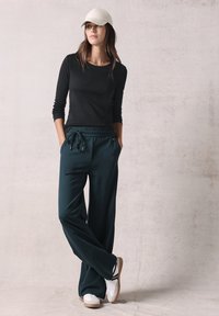 Dunkelgrüne, weite Hose mit elastischem Bund und Kordelzug. Schwarzes Langarmshirt und cremefarbene Mütze ergänzen die weißen Sneakers.