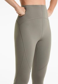 Leggings de sport taille haute en vert olive, fabriqués à partir d'un tissu lisse et extensible avec des coutures plates et des détails de panneau subtils.