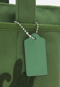 Sac en tissu texturé vert avec un motif brodé vert foncé et une étiquette en cuir verte assortie Coach New York sur une chaîne à billes argentée.
