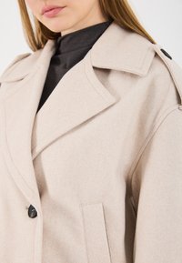 Primo piano di una donna che indossa un cappotto di lana beige su un dolcevita nero, mostrando il colletto, i dettagli delle spalle e un bottone.