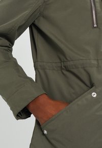 Veste légère vert olive avec une poche poitrine zippée, des poignets élastiques et des poches latérales avec fermeture à pression. Texture lisse.