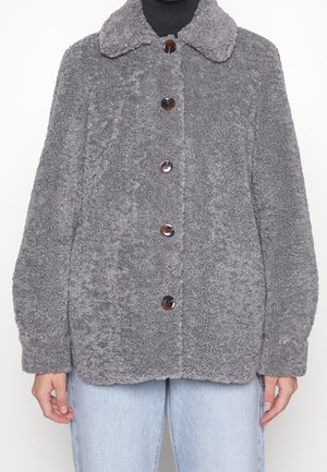 Veste grise à boutons en fausse laine avec col, portée sur un haut noir et un jean bleu clair, vue de face montrant la texture et les détails.