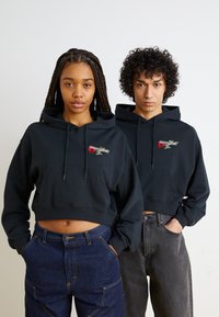 Svarte kortermede hettegensere med frontlommer har broderte roser og tekstdesign. Båret med mørk denim og grå bukser.