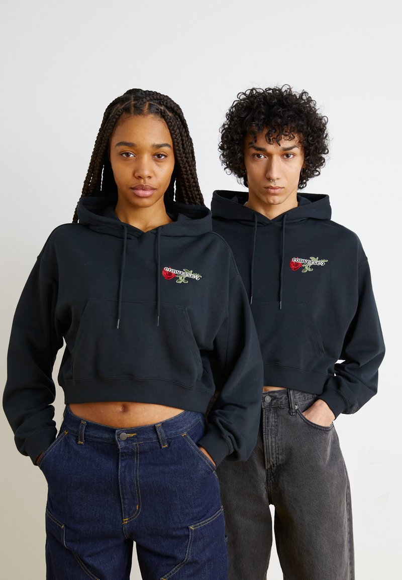 Svarte kortermede hettegensere med frontlommer har broderte roser og tekstdesign. Båret med mørk denim og grå bukser.