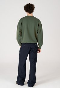 Vyras su garbanotais plaukais, dėvintis alyvuogių žalios spalvos džemperį ir tamsiai mėlynus plačius kelnes, stovi nugara į kamerą ant vienodos fono.