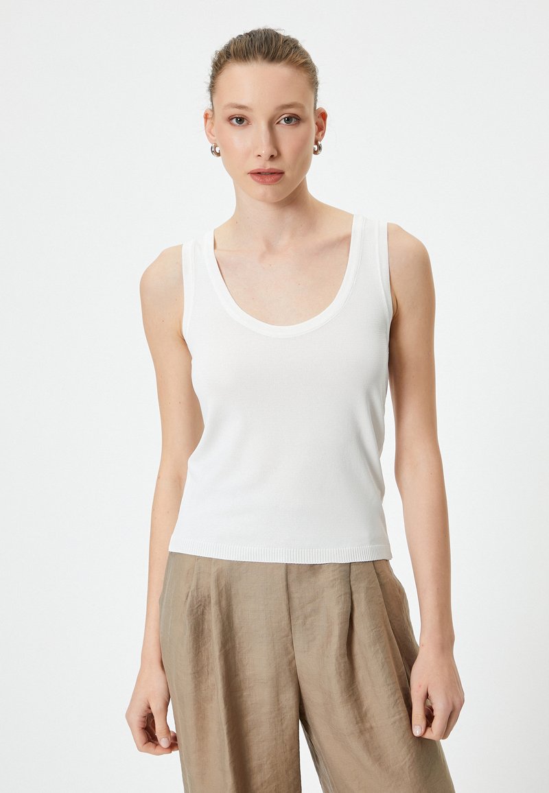 Top de tirantes ajustado en blanco con escote redondeado y tirantes anchos. Textura suave con dobladillo acanalado. Combinado con pantalones fluidos de color marrón claro.