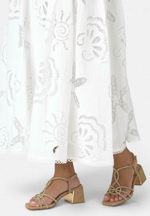 Falda blanca bordada con recortes florales combinada con sandalias de tacón grueso con tiras en color marrón claro sobre pies descalzos.