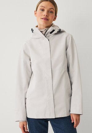 Frau trägt eine hellgraue Regenjacke mit Kapuze und Frontreißverschluss sowie blaue Jeans, stehend vor einem schlichten weißen Hintergrund.