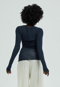 Long-sleeve navy top with a fitted shape, soft texture, and scalloped hem. Paired with light-colored, loose-fitting trousers.