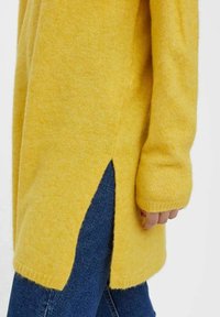 Żółty sweter z dzianiny z bocznymi rozcięciami. Miękka tekstura, długie rękawy, ściągacze przy mankietach i luźny krój. Noszony z niebieskimi dżinsami.