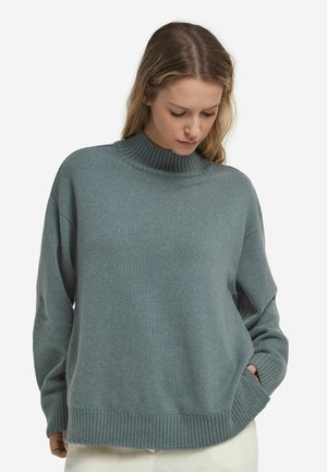 Kvinde iført en løs grå sweater med høj hals og ribstrikkede manchetter og kant, kigger ned med den ene hånd i lommen.