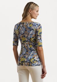 Top in maglia floreale con maniche a tre quarti, caratterizzato da un motivo blu, beige e verde. Silhouette aderente e orlo leggermente curvo.