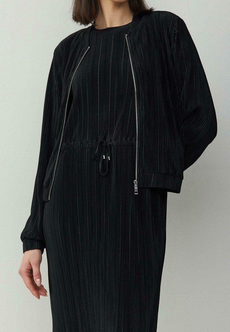 Femme portant une robe plissée noire avec une veste bomber noire assortie et zippée, debout devant un fond clair uni.