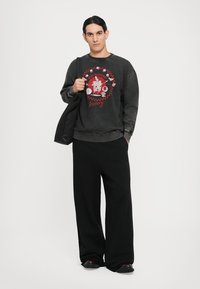 Junger Mann mit schwarzen Haaren trägt einen dunklen, übergroßen Sweatshirt mit roter Grafik, weite schwarze Hosen, schwarze Schuhe und trägt eine schwarze Schultertasche.