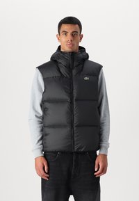 Chaleco acolchado negro con cuello alto y capucha, cierre de cremallera y detalle del logo verde. Lleva puesto sobre una camiseta de manga larga gris.