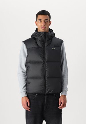Zwarte gewatteerde vest met een hoge kraag en capuchon, ritssluiting en groen logo-detail. Draag over een grijze lange mouwen shirt.
