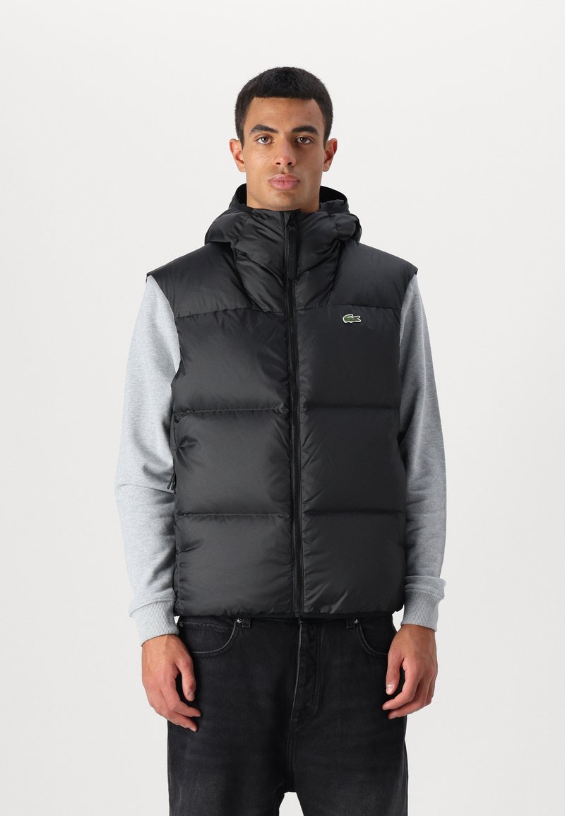 Chaleco acolchado negro con cuello alto y capucha, cierre de cremallera y detalle del logo verde. Lleva puesto sobre una camiseta de manga larga gris.