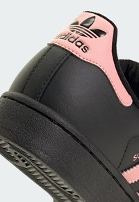 Μαύρο δερμάτινο αθλητικό παπούτσι με ροζ λεπτομέρεια κοντά στον αστράγαλο, υφασμάτινη σόλα και λείος επάνω με σήμα Adidas.