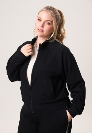 Vrouw met blond paardenstaart, draagt een zwart jack met ritssluiting over een licht shirt, één hand die de kraag aanpast, neutrale achtergrond.