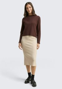 Brązowy sweter z okrągłym dekoltem, połączony z kremową spódnicą midi. Modelka ma na sobie czarne buty na platformie i ciemne skarpetki. Gładkie faktury, dopasowana sylwetka.