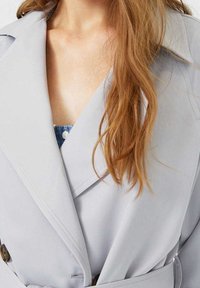 Zbliżenie na osobę w jasnoszarym płaszczu trenchowym z paskiem, noszącą niebieską odzież z guzikami, z długimi falującymi rudobrązowymi włosami widocznymi.