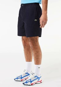 Pantaloni corti blu navy con vita elastica e tasche laterali, caratterizzati da un piccolo logo verde Lacoste. Indossati con calzini bianchi e scarpe da ginnastica multicolore.