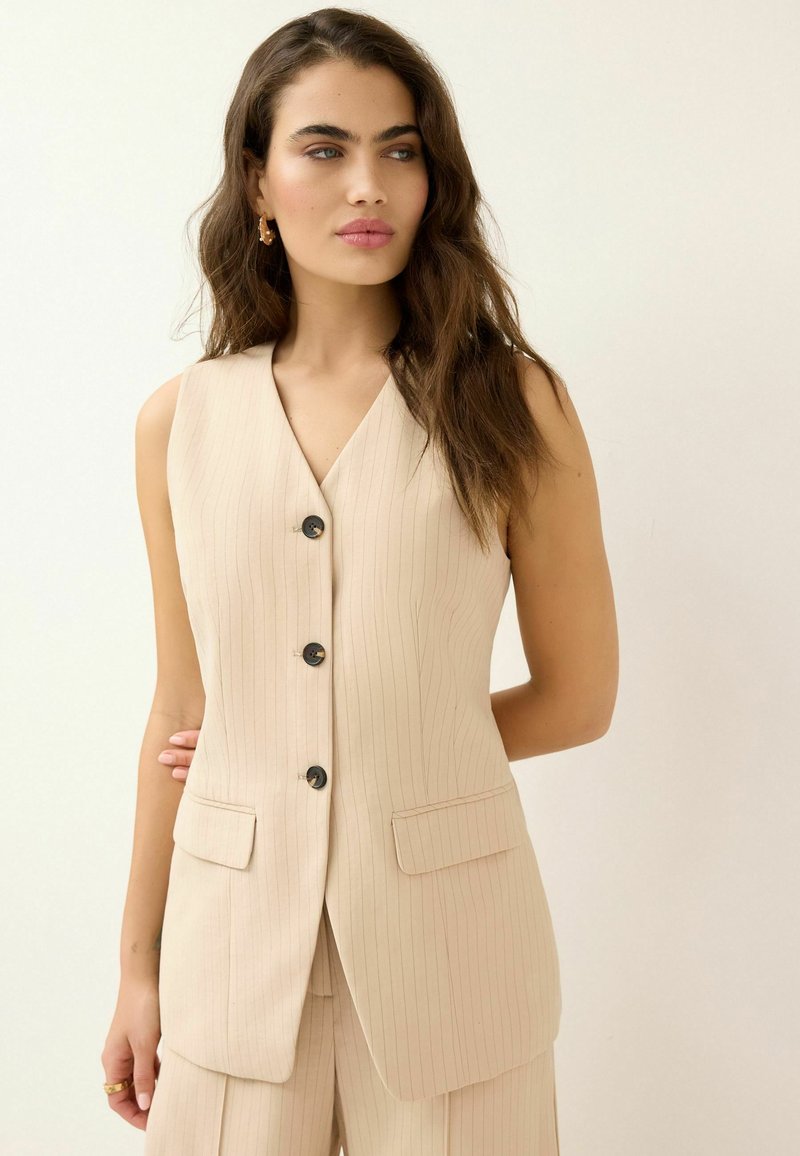 Gilet beige à rayures verticales avec un décolleté en V, des boutons noirs et deux poches avant. Le tissu semble lisse avec une coupe ajustée.
