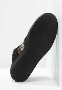 Sneaker noir avec une semelle en caoutchouc texturée comportant des lettres en relief. Accents en suède marron sur les côtés et une bande de logo blanche.